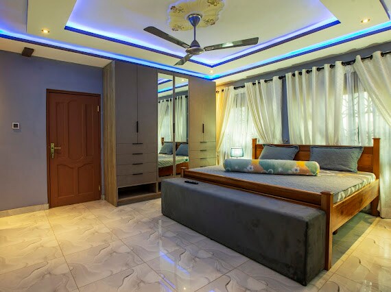 A spacious bedroom features a large bed with a padded base, complemented by decorative cushions. Soft light streams through sheer curtains, and a ceiling fan provides air circulation. A sliding door wardrobe with mirrored panels adds functionality, enhancing the overall serene atmosphere.