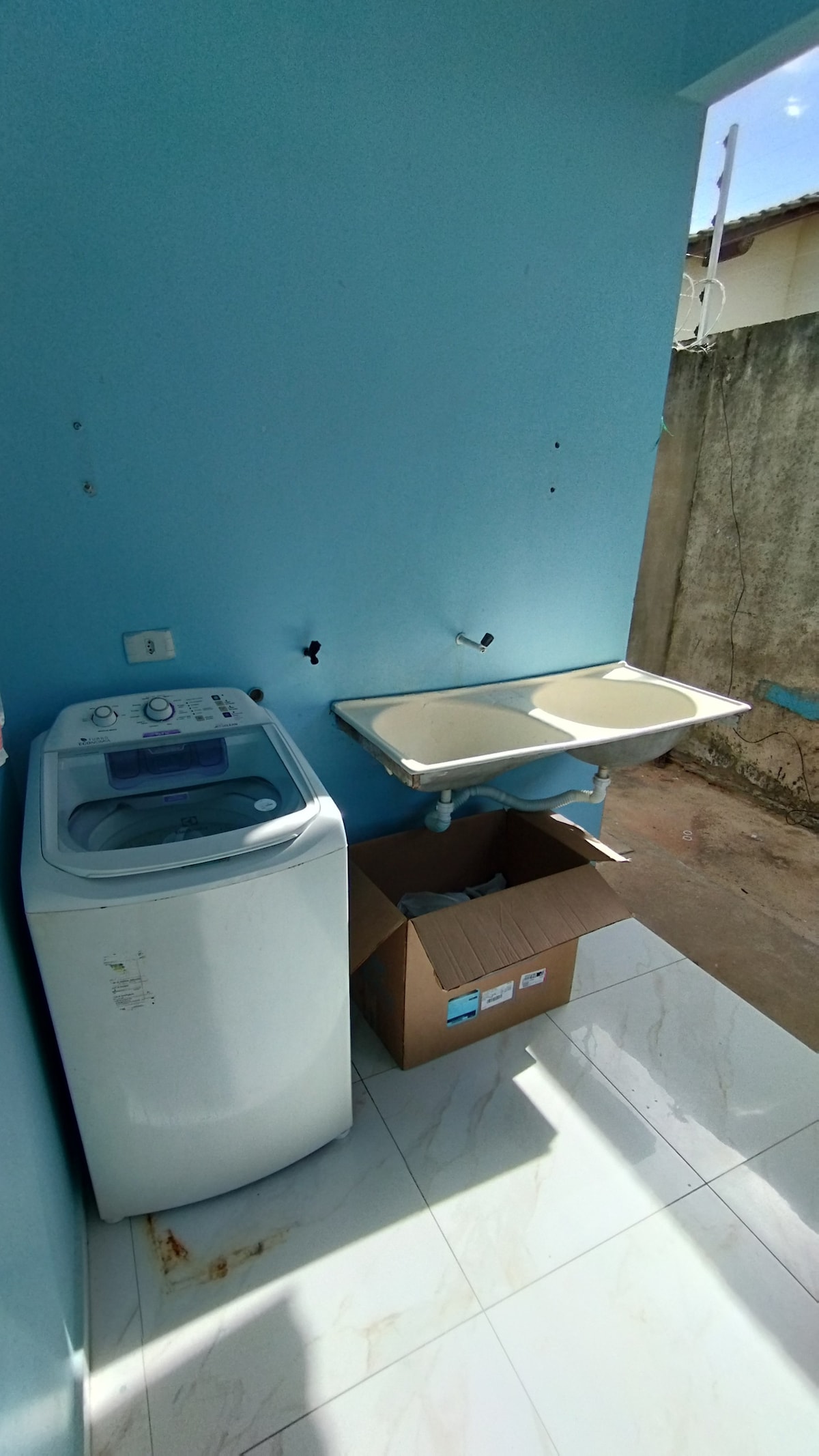 A laundry area is featured with a washing machine positioned beside a light-colored sink with countertop space. The walls are painted in a soft blue, and the floor presents white tiles that reflect natural light. A cardboard box sits nearby.