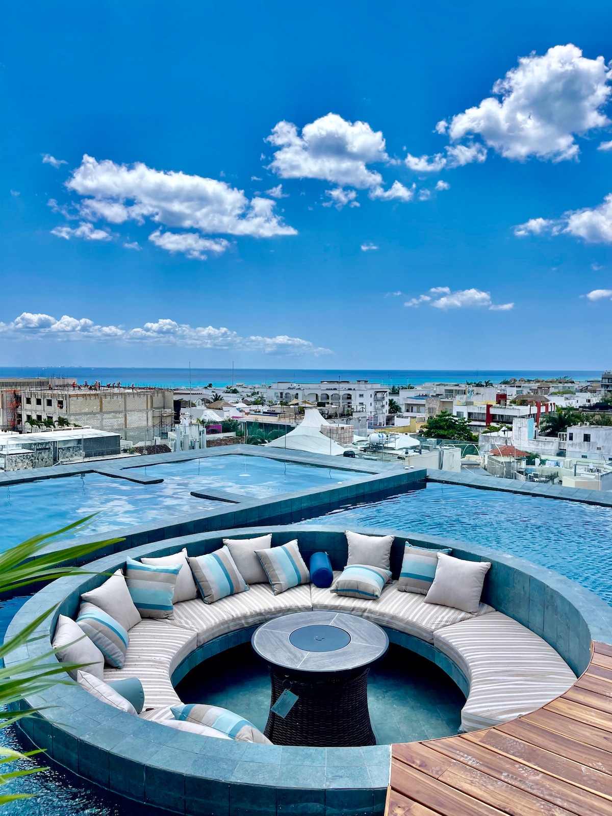 A spacious circular seating area is surrounded by a tranquil pool, offering a panoramic view of the coastal landscape. Light blue cushions are arranged around a central table, creating a relaxing space for gatherings, with the ocean visible in the background under a clear blue sky.
