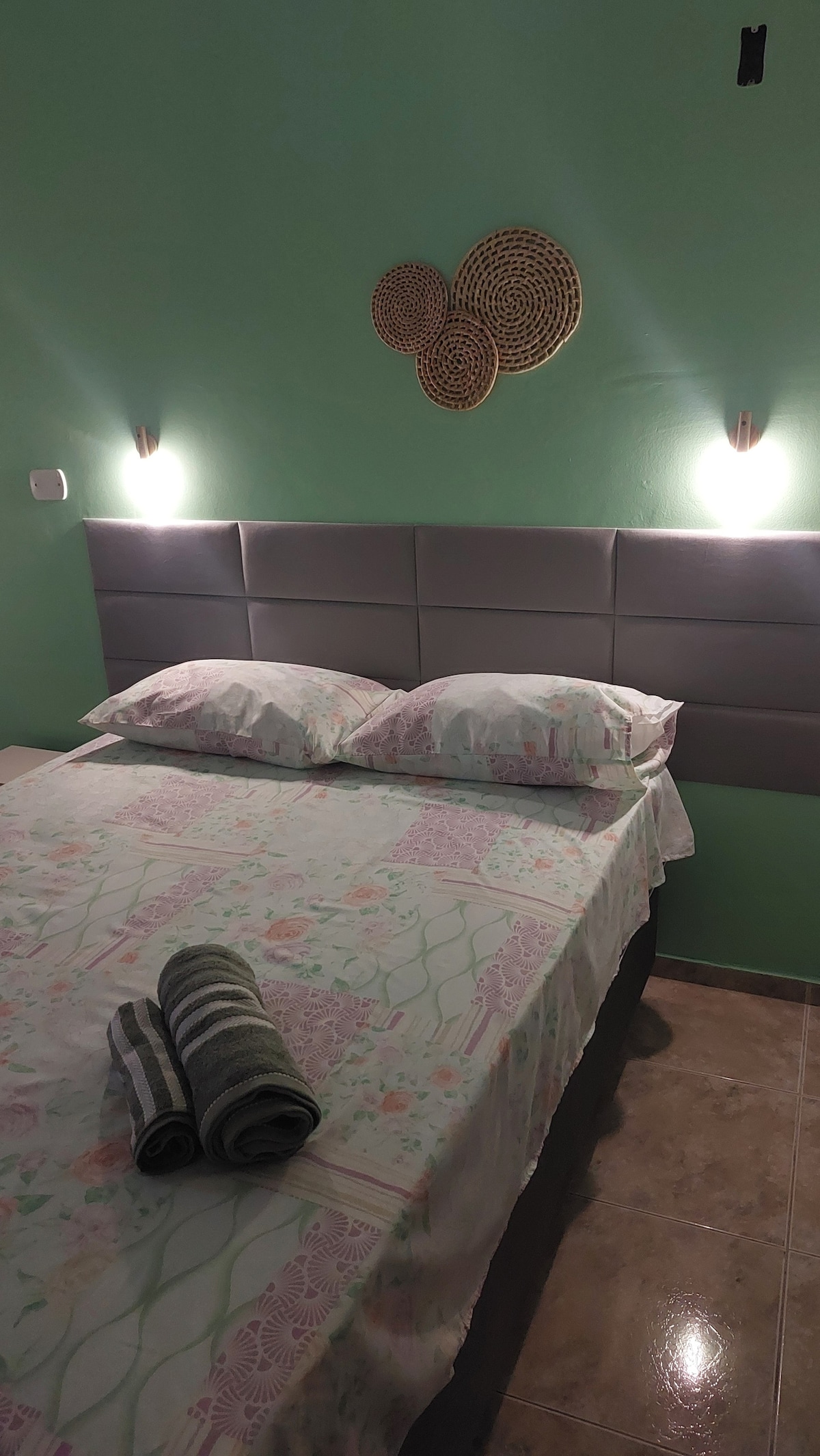 A cozy sleeping area features a spacious bed with a floral-patterned quilt. Two soft pillows rest against a padded headboard. Rolled towels are placed neatly on the bed. Soft lighting from wall-mounted fixtures highlights the calming green walls.