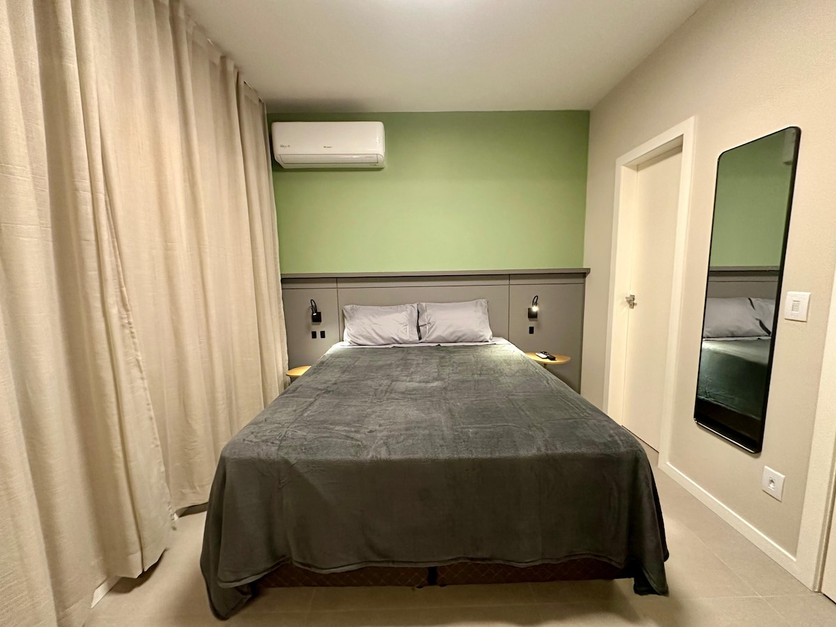 A spacious bed is dressed in a dark gray coverlet, complemented by light gray pillows. Bedside lamps are positioned on either side, while a large mirror reflects the room. A subtle green accent wall enhances the calming ambiance, and light-colored curtains frame a window.