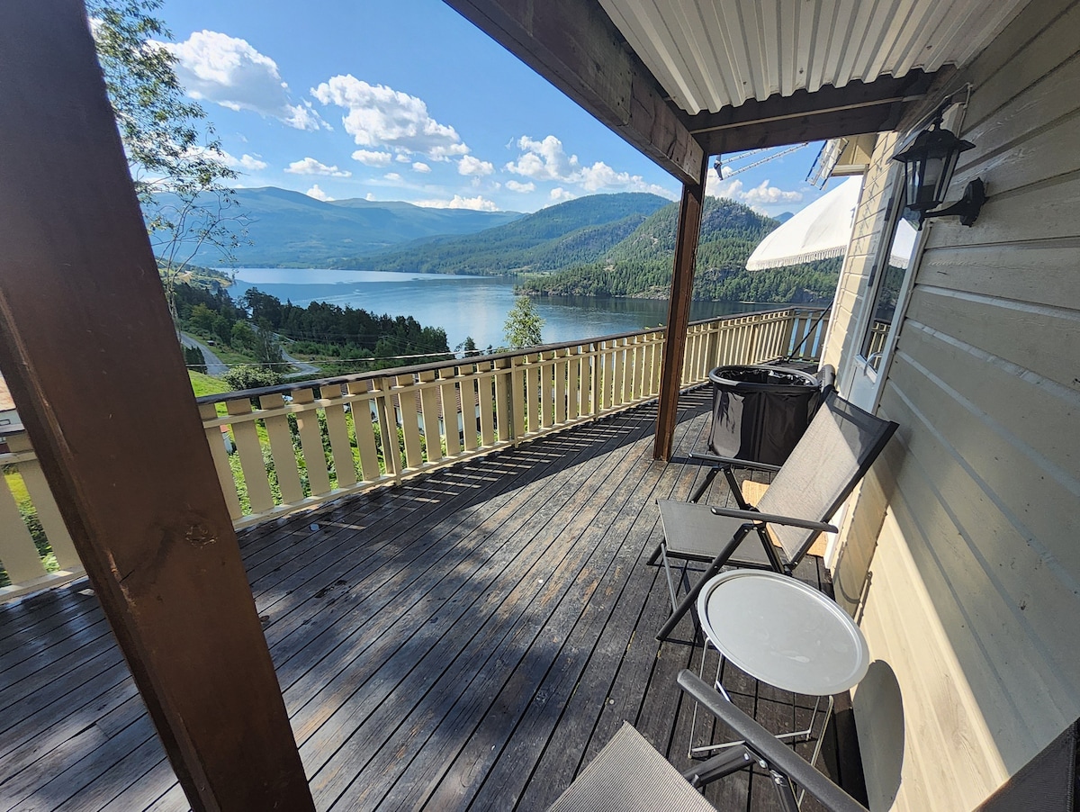 A spacious deck offers a view of the tranquil body of water surrounded by hills and trees. Two chairs and a small table are positioned on the wooden surface, while a hot tub is partially visible, creating an inviting outdoor space for relaxation.