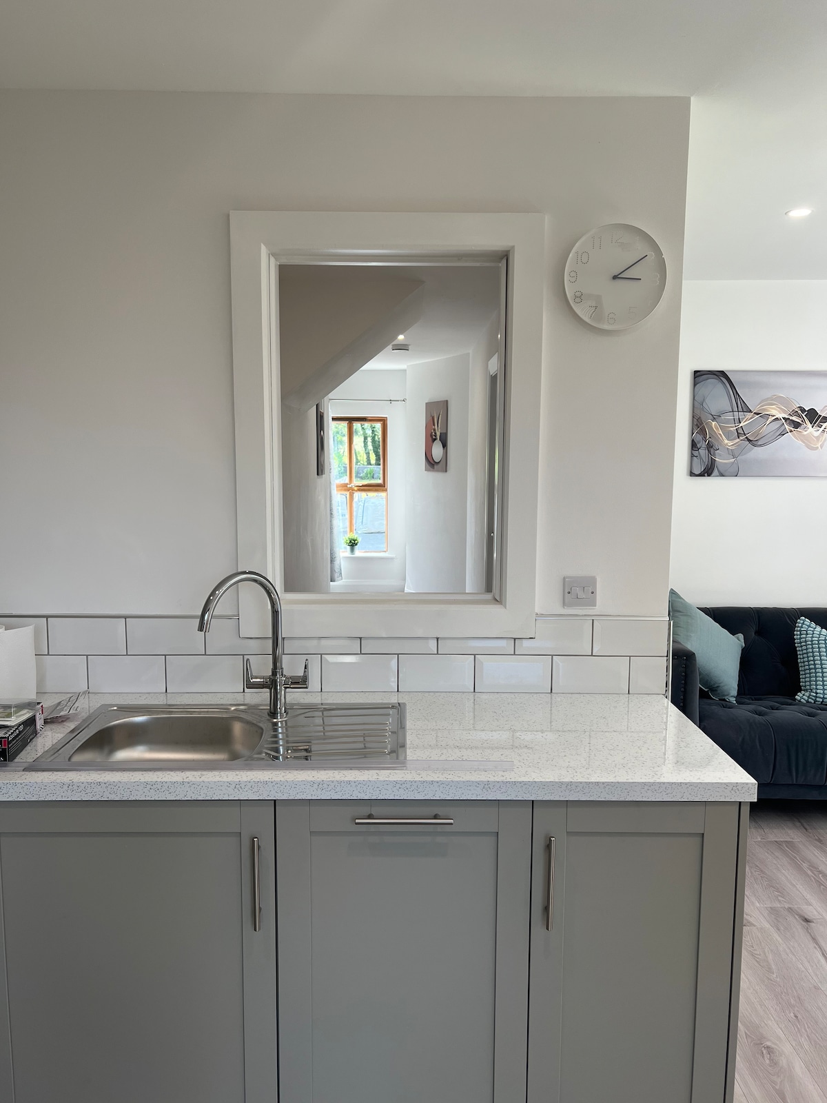The modern kitchen features sleek gray cabinetry and a stainless steel sink. A large mirror reflects natural light, enhancing the bright atmosphere. A wall clock and a polished countertop add functional elements, while a glimpse of the living area is visible in the background.