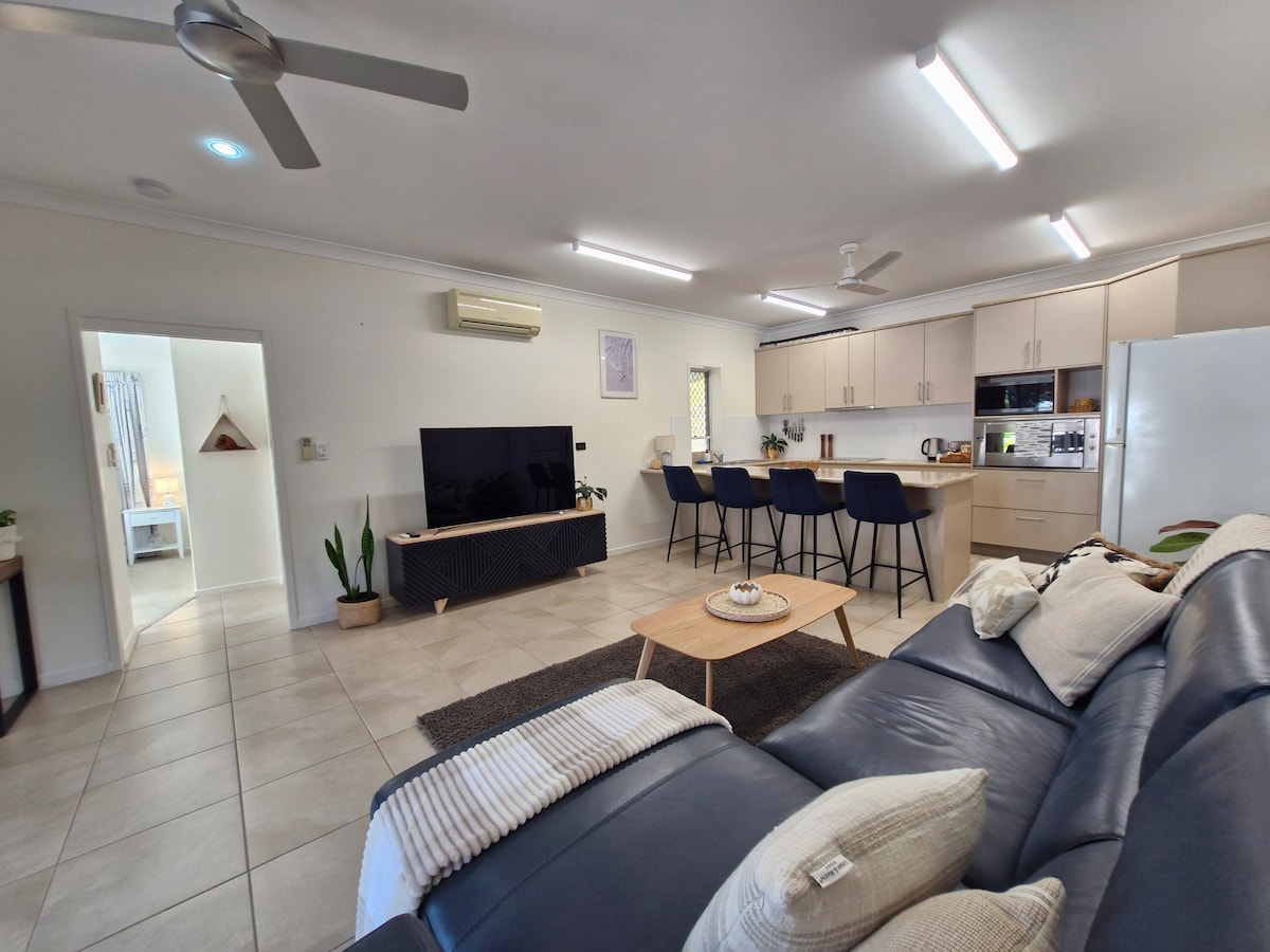 A spacious living area is visible, featuring a comfortable sectional sofa and a minimalist coffee table. A large flat-screen television is positioned against the wall, accompanied by several barstools around a kitchen island. Natural light enters through the windows, enhancing the neutral decor.
