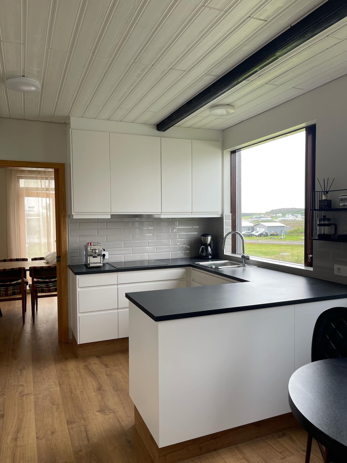 A modern kitchen features sleek white cabinetry and a dark countertop. Natural light floods the space through a large window, revealing views of the outdoors. Equipped with essential appliances, the kitchen layout promotes functionality and ease for meal preparation.