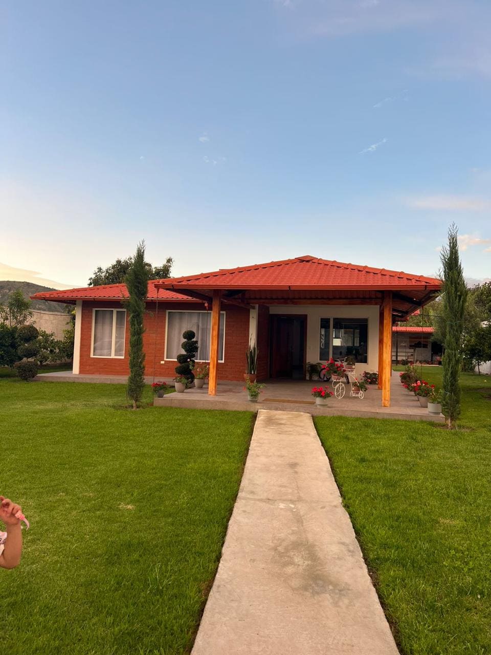 A charming house features a red-tiled roof and warm wooden beams, surrounded by neatly trimmed green grass. Colorful flower beds are positioned alongside the entrance, while a stone path leads to the front door. Lush trees are visible in the background, enhancing the natural setting.