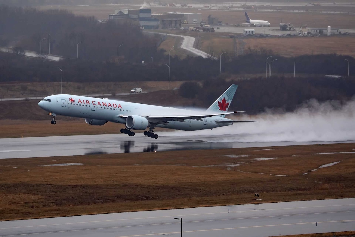 A large aircraft from Air Canada is captured during takeoff on a wet runway. A mist of water is created as the plane accelerates, with a blurred airport landscape visible in the background under a cloudy sky.