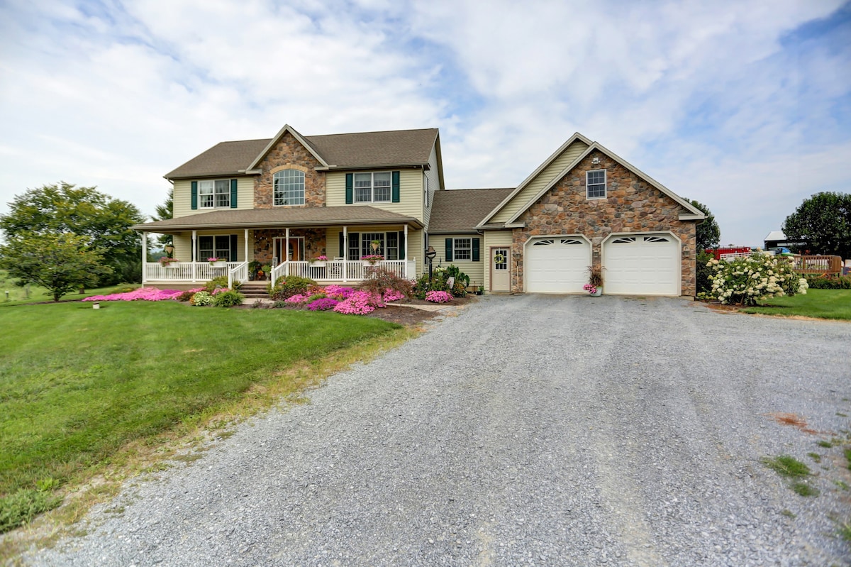 A two-story house is shown with a stone façade, flanked by a well-maintained gravel driveway and vibrant flower beds. The wide front porch is accessible, and large windows allow natural light to fill the interior. Green grass surrounds the property, contributing to the countryside setting.