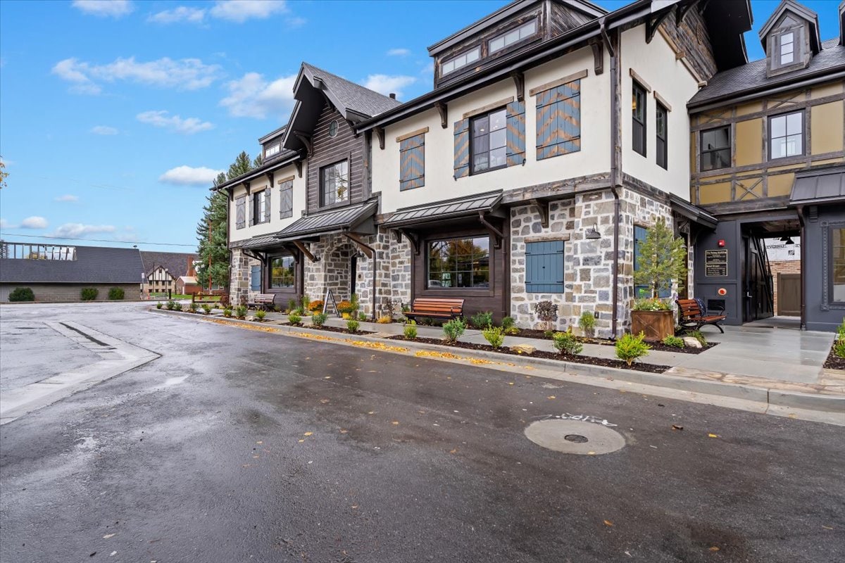 A modern building exterior is showcased with a combination of stone and wood siding. Large windows provide light while benches are placed along the front, surrounded by neatly arranged shrubs. The paved area reflects recent rainfall, enhancing the clean and inviting look of the entrance.