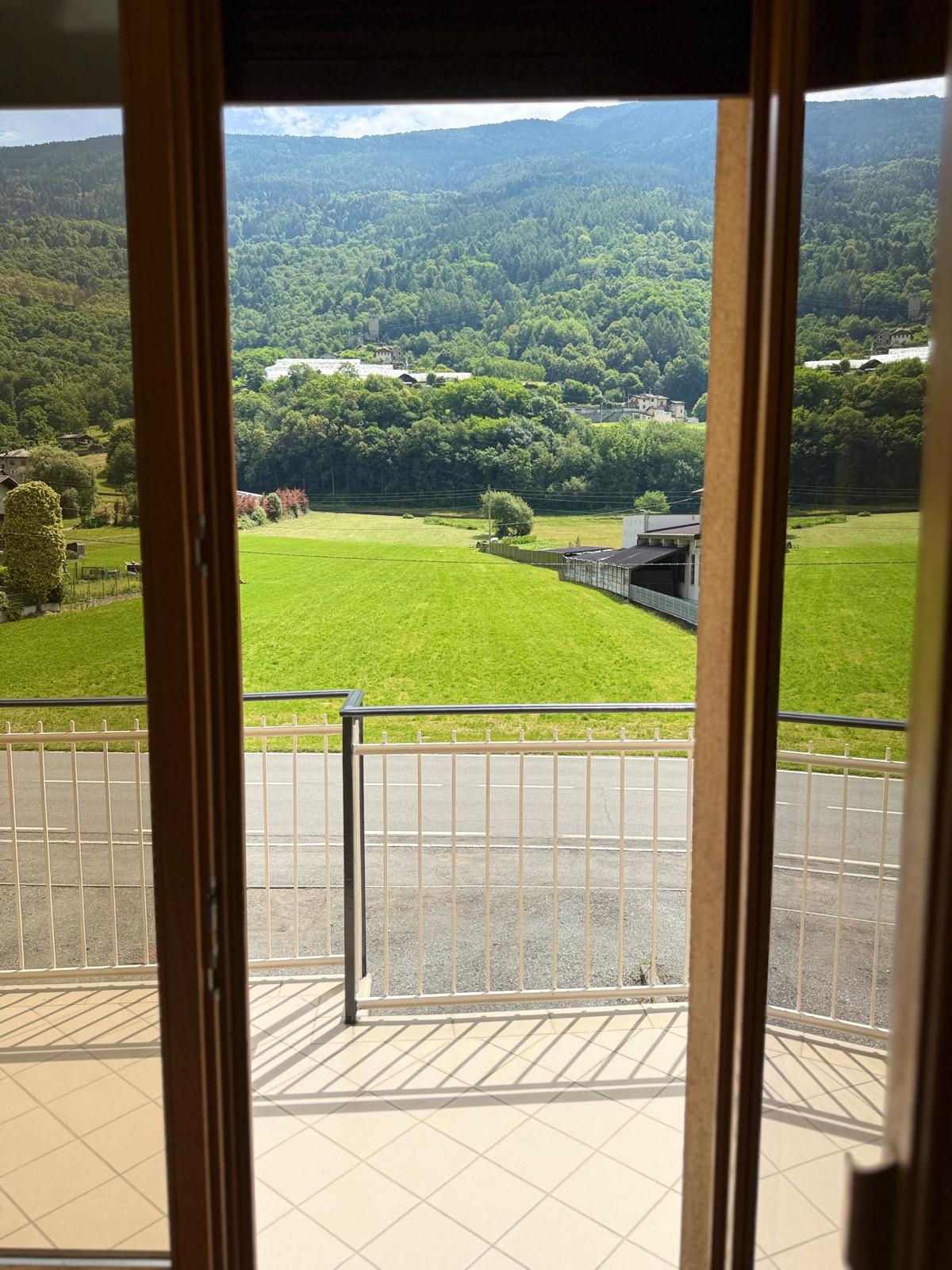 A balcony view is presented, framed by wooden doors opening to a lush green landscape with rolling hills. Natural light streams in, highlighting the tiled floor. The distant mountains serve as a serene backdrop, with trees and fields extending into the horizon.