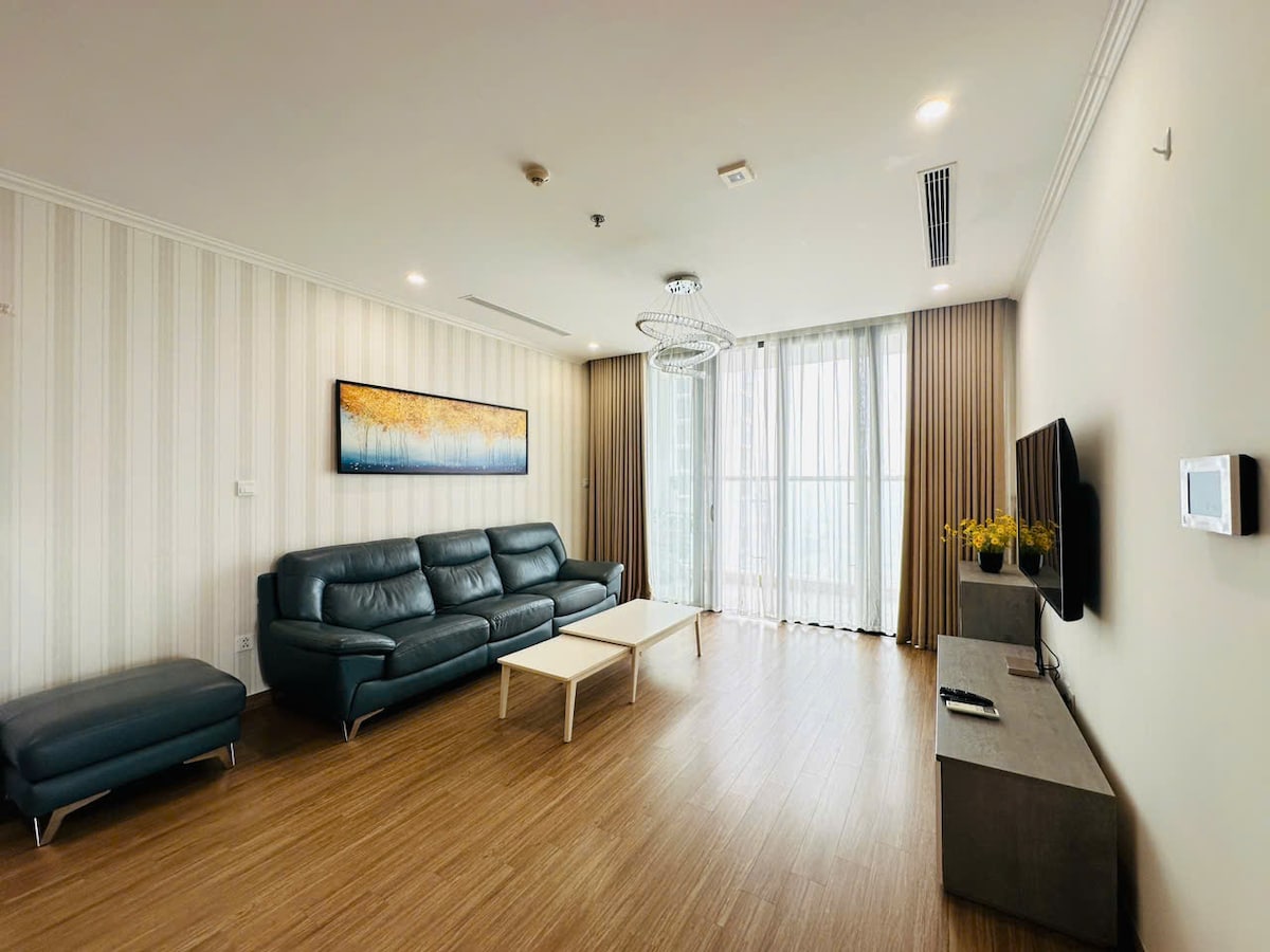 A modern living area features a dark leather sofa set paired with a light coffee table. Large windows allow natural light to fill the space, while elegant curtains frame the view. A flat-screen TV is mounted on a neutral-toned wall unit, creating a sleek design.