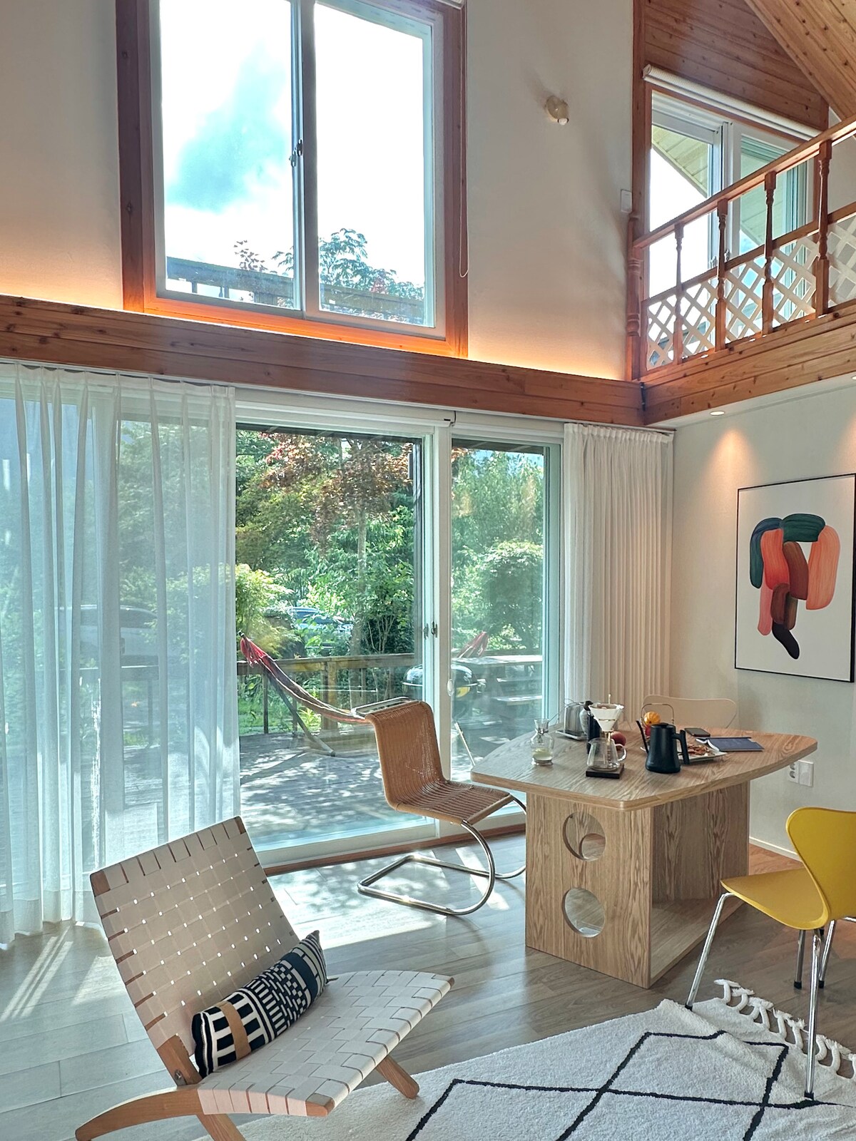 A bright living space is highlighted by large windows allowing natural light to fill the area. The decor includes a wooden table set with a coffee maker and cups, complemented by stylish seating arrangements. A view of greenery is visible through sheer curtains.