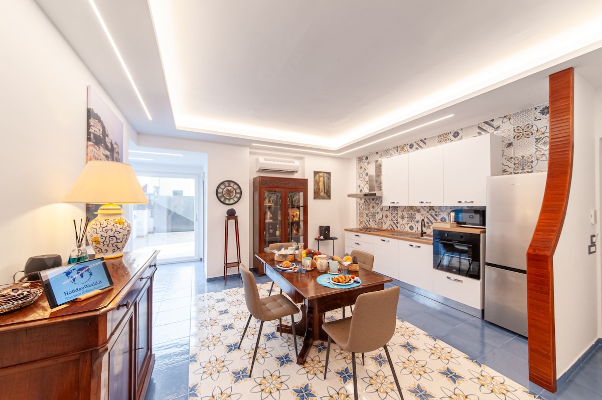 An inviting dining space is presented with a wooden table set for a meal, featuring assorted dishes and drinks. Bright blue tile flooring complements the white cabinetry and patterned backsplash in the kitchen. Natural light filters in from a nearby door, enhancing the cheerful atmosphere.