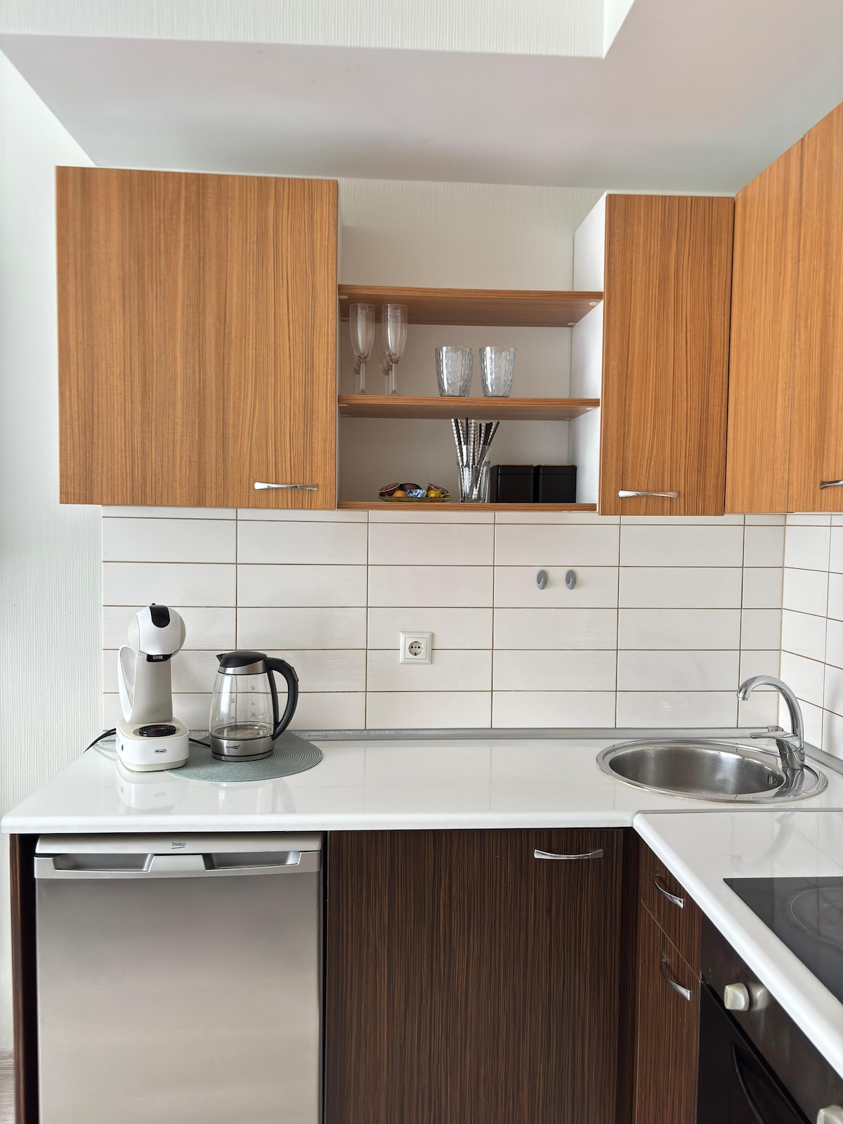 A modern kitchen is presented with wooden cabinets and white tiled backsplash. An electric kettle and coffee maker are placed on the counter, alongside a stainless-steel sink. Glassware is displayed on a shelf above, and a dishwasher is integrated into the cabinetry.