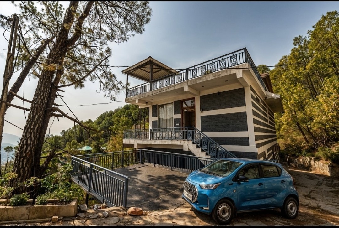 The exterior of a spacious 4BHK apartment is showcased, surrounded by lush greenery. A small blue car is parked in the driveway, which features a flat, paved surface. The building is designed with modern architecture and has a welcoming, accessible entrance, promoting ease of access.