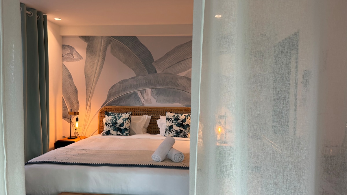A serene sleeping area features a spacious bed adorned with decorative pillows. The background showcases a mural of large leaves, enhancing the calming atmosphere. Soft curtains partially frame the scene, allowing natural light to filter into the space.