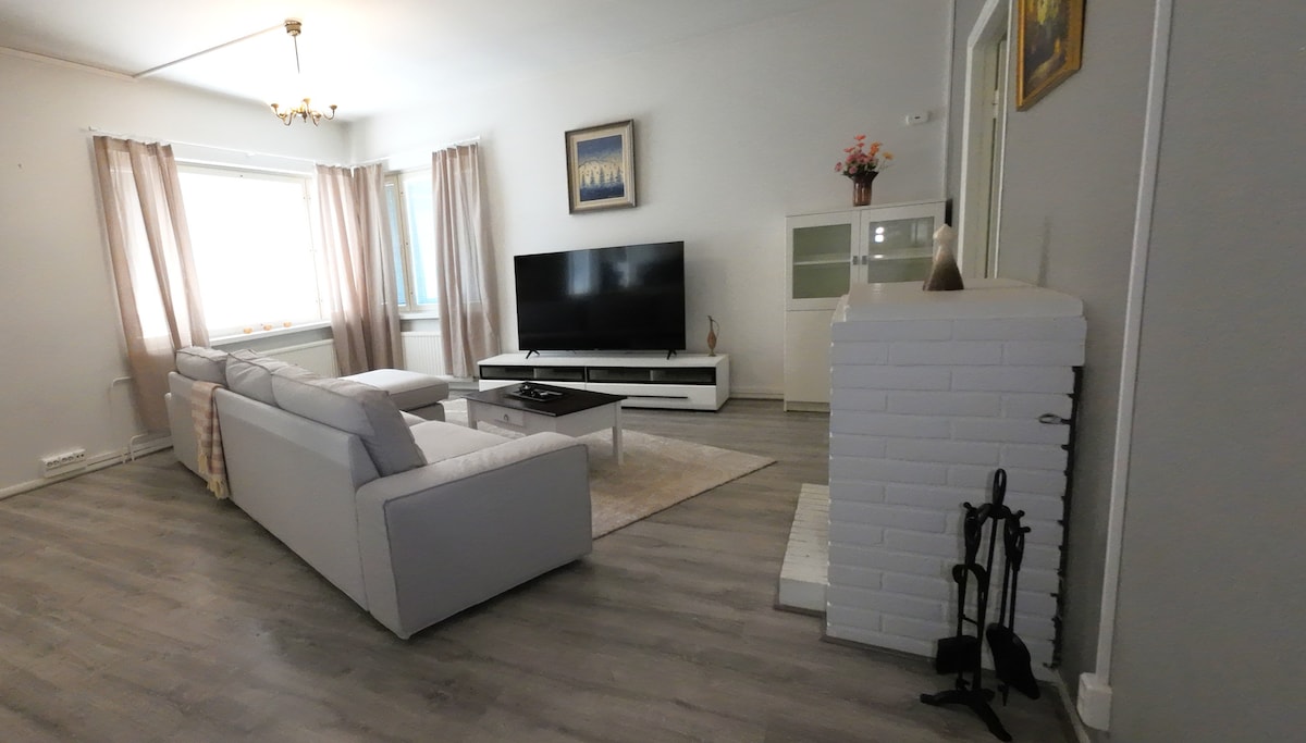 The living room features a light-colored sectional sofa positioned to face a large television. Soft natural light fills the space through sheer curtains adorning the window. A white brick fireplace adds a touch of warmth and charm to the inviting atmosphere.