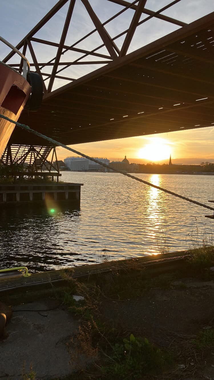 A golden sunset is captured behind a silhouetted structure with a bridge overhead. The calm water reflects the warm light, while the distant skyline, featuring church steeples, adds depth to the image. Vegetation grows along the waterfront, enhancing the natural setting.