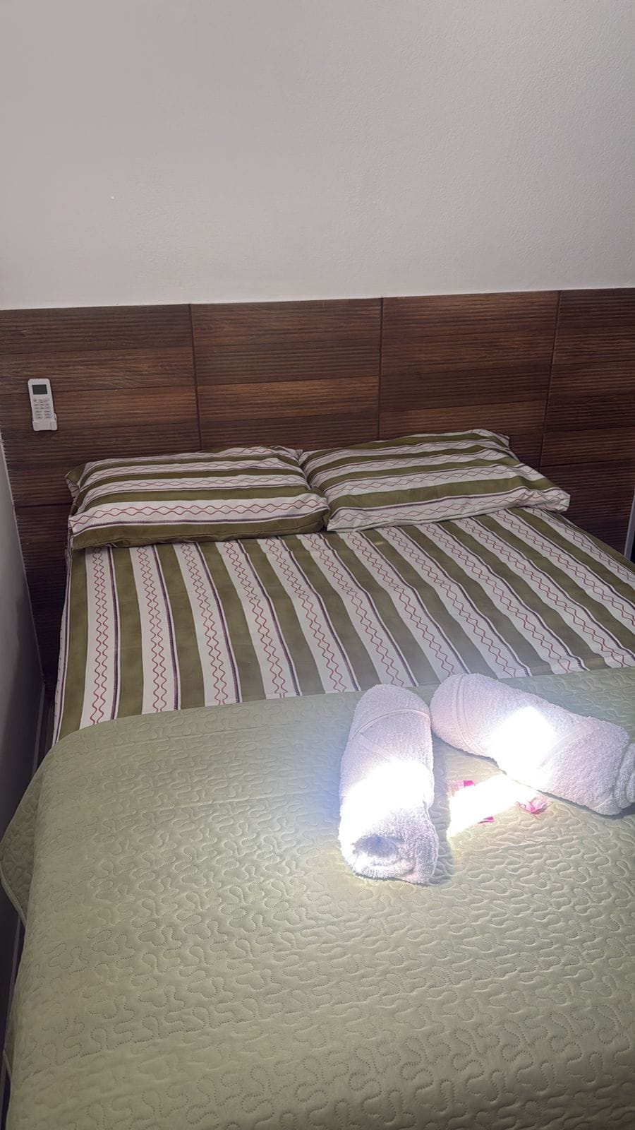 A spacious bed is set against a wooden headboard, fitted with striped green and white bedding. Two pillows add comfort, while neatly rolled towels are placed at the foot of the bed. Natural light floods the space, enhancing its clean and inviting appearance.