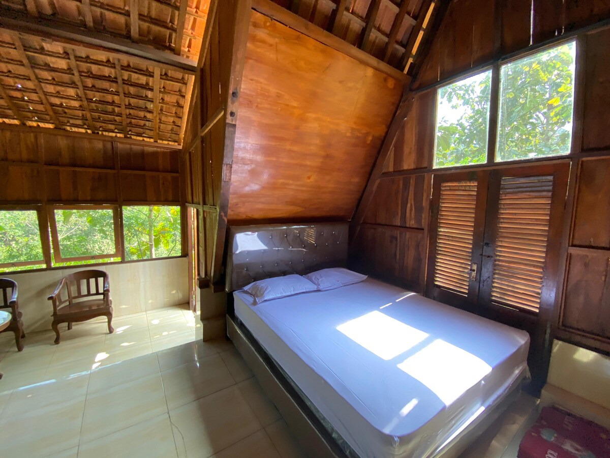 A cozy bedroom is framed by large windows allowing natural light to pour in, highlighting the wood-paneled walls and ceiling. The spacious bed is dressed in white linens, and two wooden chairs are positioned nearby, offering a serene space for relaxation.
