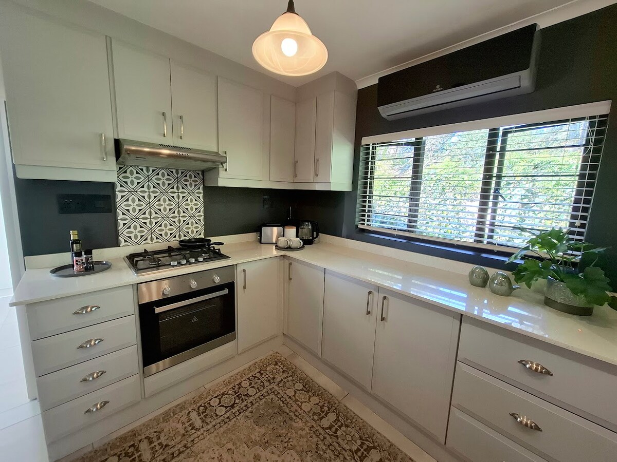 A modern kitchen features sleek white cabinetry and a stylish backsplash with geometric patterns. Stainless steel appliances are positioned along the countertop, which is complemented by a subtle area rug. Natural light filters through the window, highlighting potted plants on the sill.