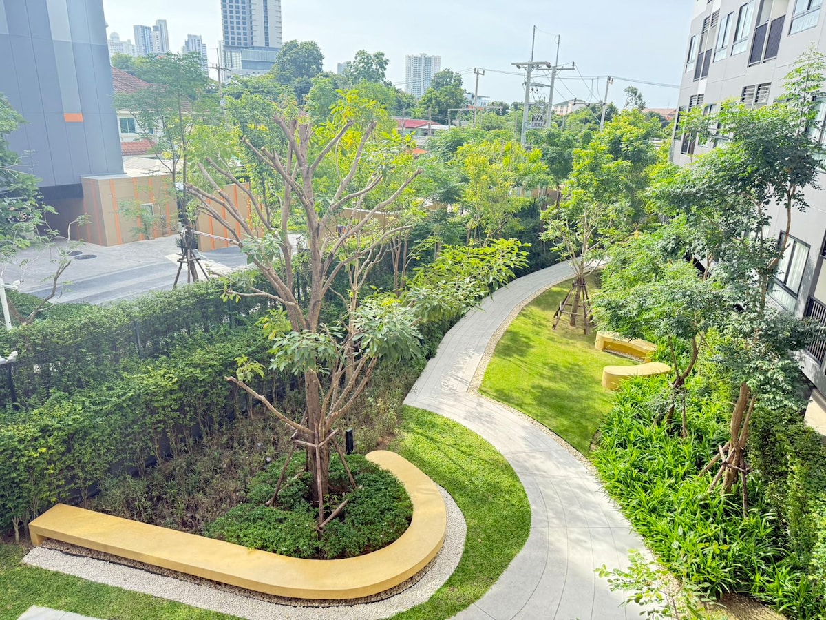 The image displays a landscaped outdoor area featuring curving pathways bordered by lush greenery. Mature trees provide shade, while benches are positioned along the path, inviting relaxation. The background includes modern buildings and a few power lines, enhancing the urban feel of the surroundings.