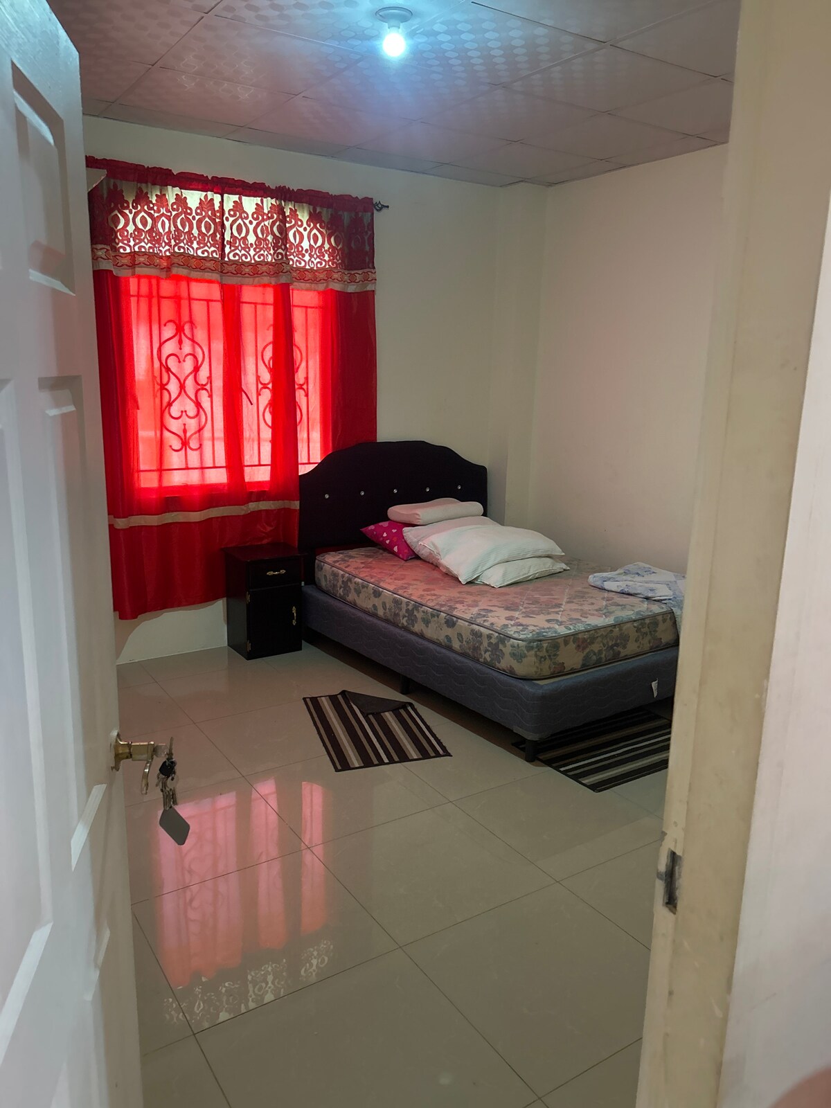 A small bedroom is presented with a single bed, adorned with soft pillows and a patterned blanket. Bright red curtains frame the window, allowing natural light to illuminate the space. A dark nightstand and striped area rugs add functional elements to the room.