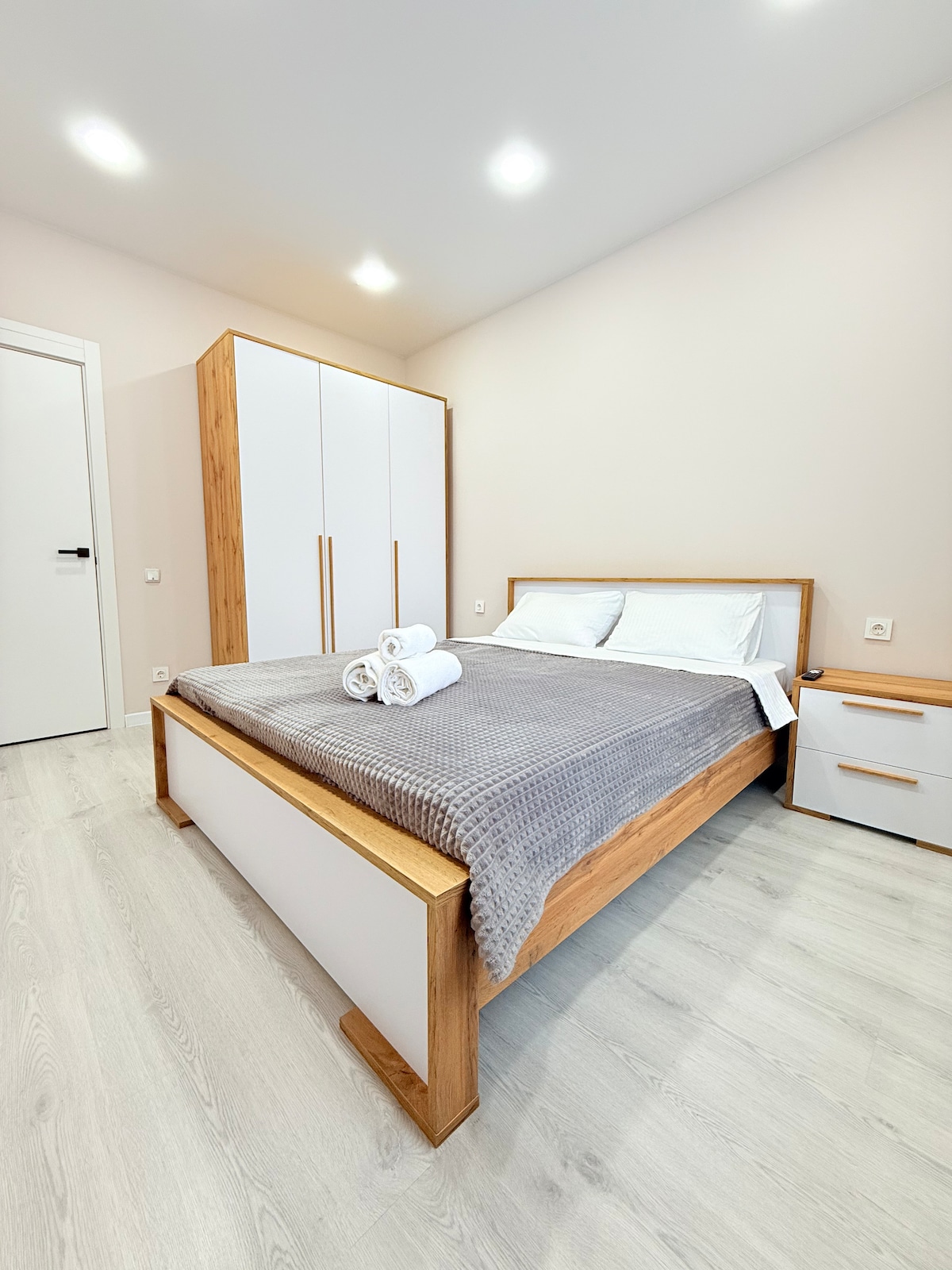 A bedroom is presented, featuring a spacious bed with light-colored linens and a soft gray throw. A modern wardrobe stands against the wall, and a bedside table is positioned beside the bed. Soft lighting enhances the neutral color scheme of the room.