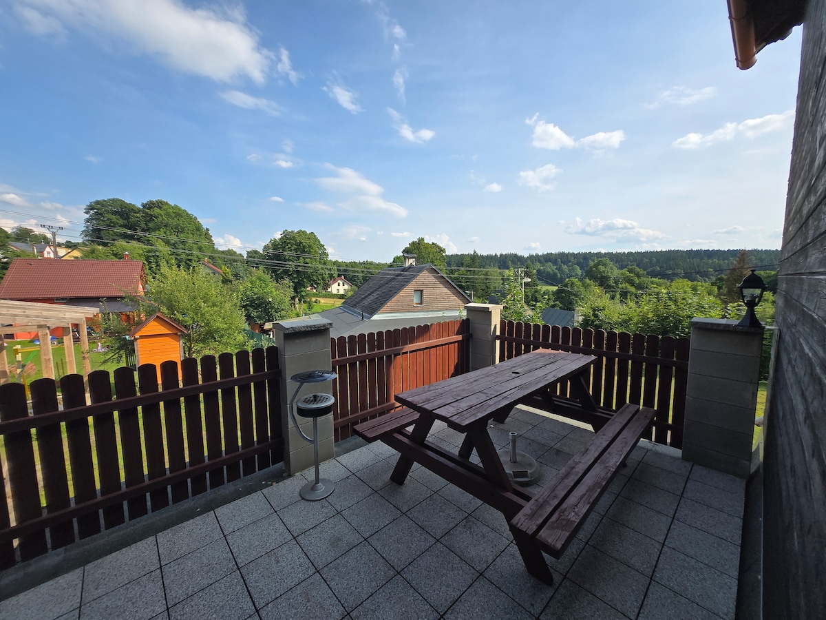 A patio area is presented, featuring a wooden picnic table and a charcoal grill. The space is surrounded by a low wooden fence, offering views of the greenery beyond. Nearby structures are visible, including a play area and shaded trees under a clear blue sky.
