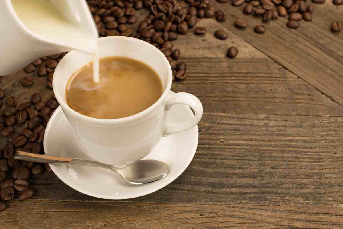 A white cup of coffee sits on a matching saucer, surrounded by scattered coffee beans on a rustic wooden surface. A silver spoon rests beside the cup, while a stream of milk is being poured into the coffee.