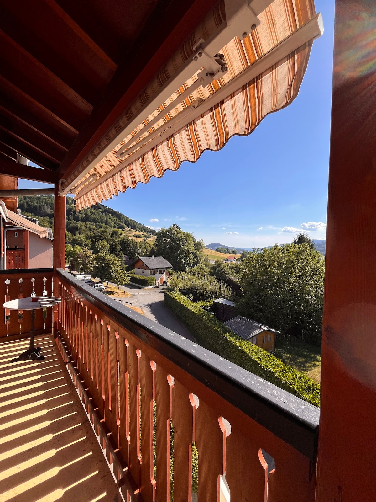 A balcony view showcases a lush valley and neighboring houses, framed by wooden railings. The awning offers shade from the sunlight, while the surrounding greenery and hills create a serene backdrop. A clear sky with a hint of clouds enhances the inviting outdoor space.