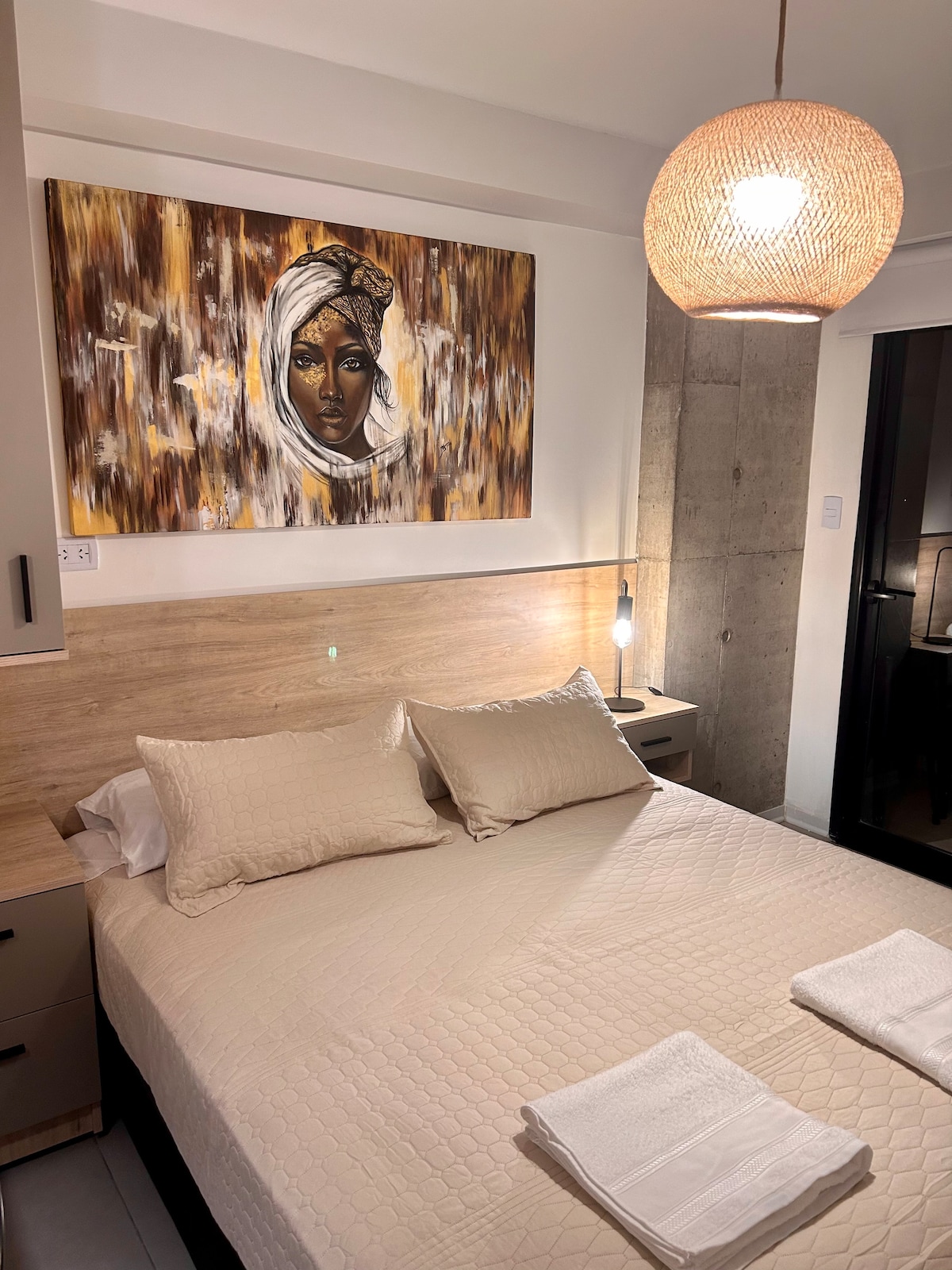 A bedroom features a spacious bed with neutral linens and two folded towels. Above the bed, an abstract artwork adds visual interest, while a modern pendant light provides soft illumination. A sliding door leads to a balcony, enhancing natural light in the room.