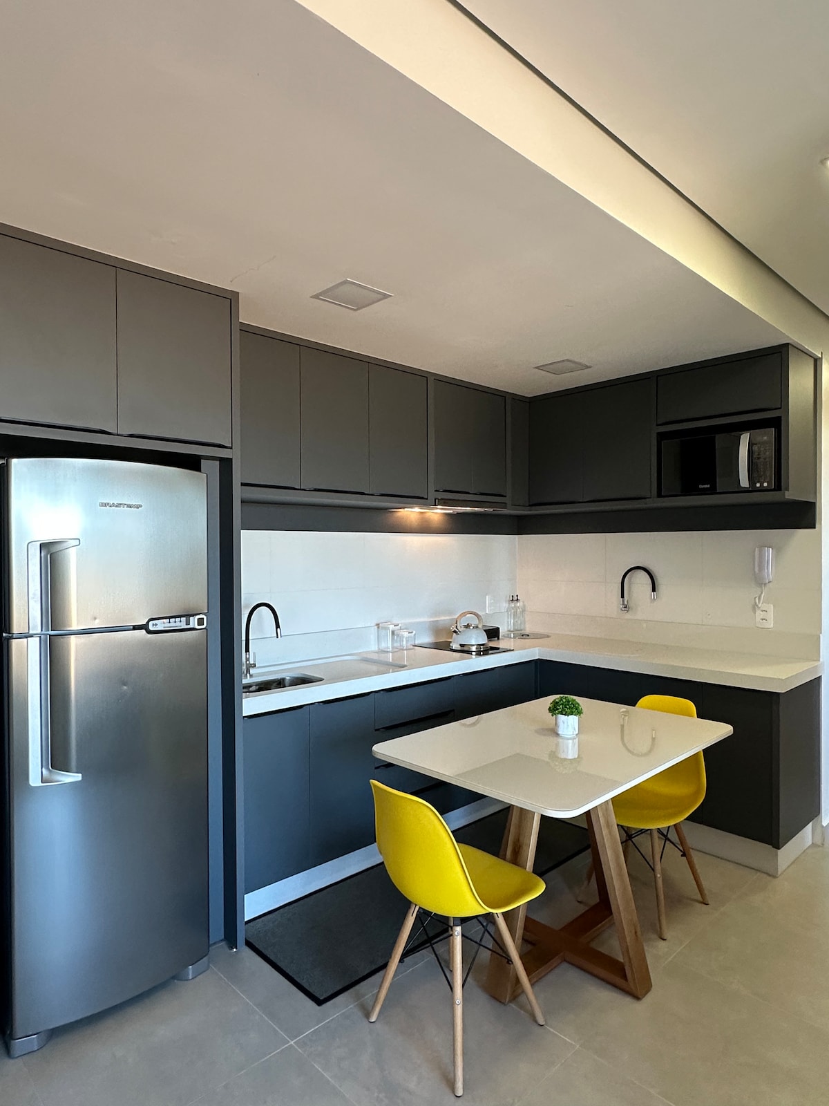 A modern kitchen area features sleek gray cabinetry and a stainless steel refrigerator. A compact dining table with yellow chairs is positioned in the center. Counter space is equipped with a microwave and kitchen sink, creating a practical setup for meal preparation.