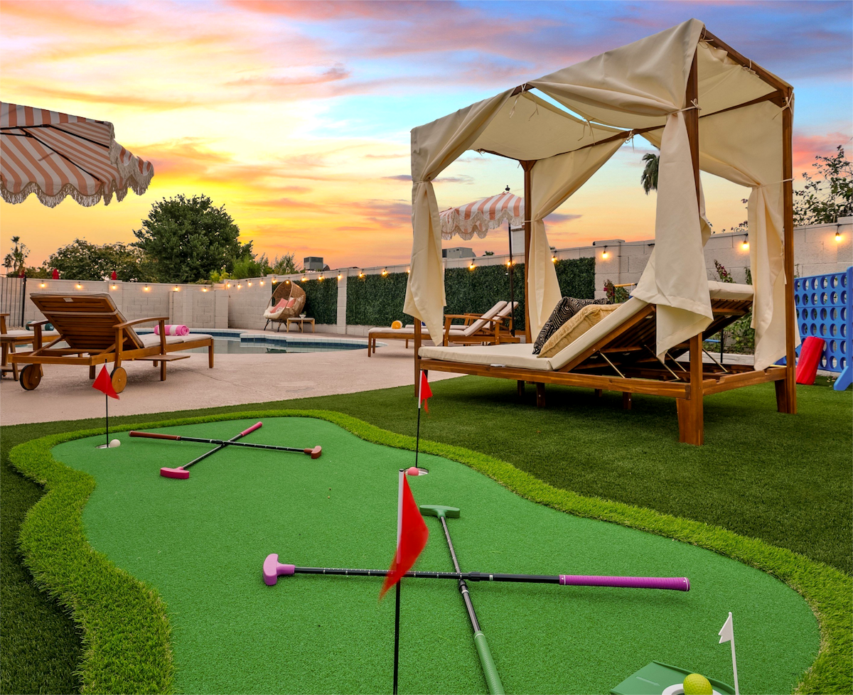 A private mini golf area features a vibrant green synthetic putting surface, adorned with colorful flags. A shaded lounge area with a canopy provides a comfortable retreat, while wooden lounge chairs invite relaxation. The sky displays a warm sunset, creating a serene setting.