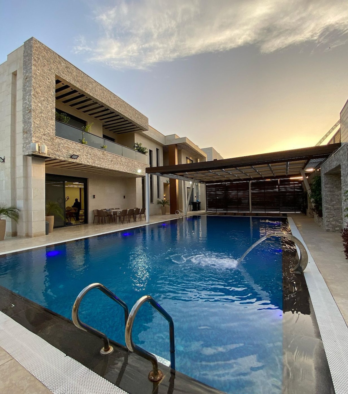 A modern villa features a large pool with water features, surrounded by sleek stone architecture. Multiple lounge areas are positioned around the pool, and outdoor seating is available under a shaded patio. The scene is complemented by a sunset sky.