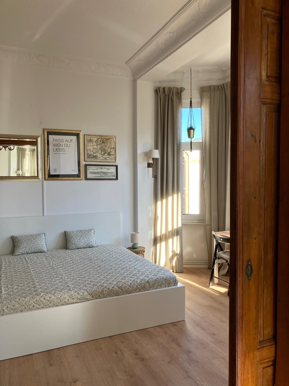A bright bedroom features a spacious bed covered with a textured blanket, accompanied by two decorative pillows. Wall art and framed prints add character to the room, while natural light streams in through a window adorned with light curtains.