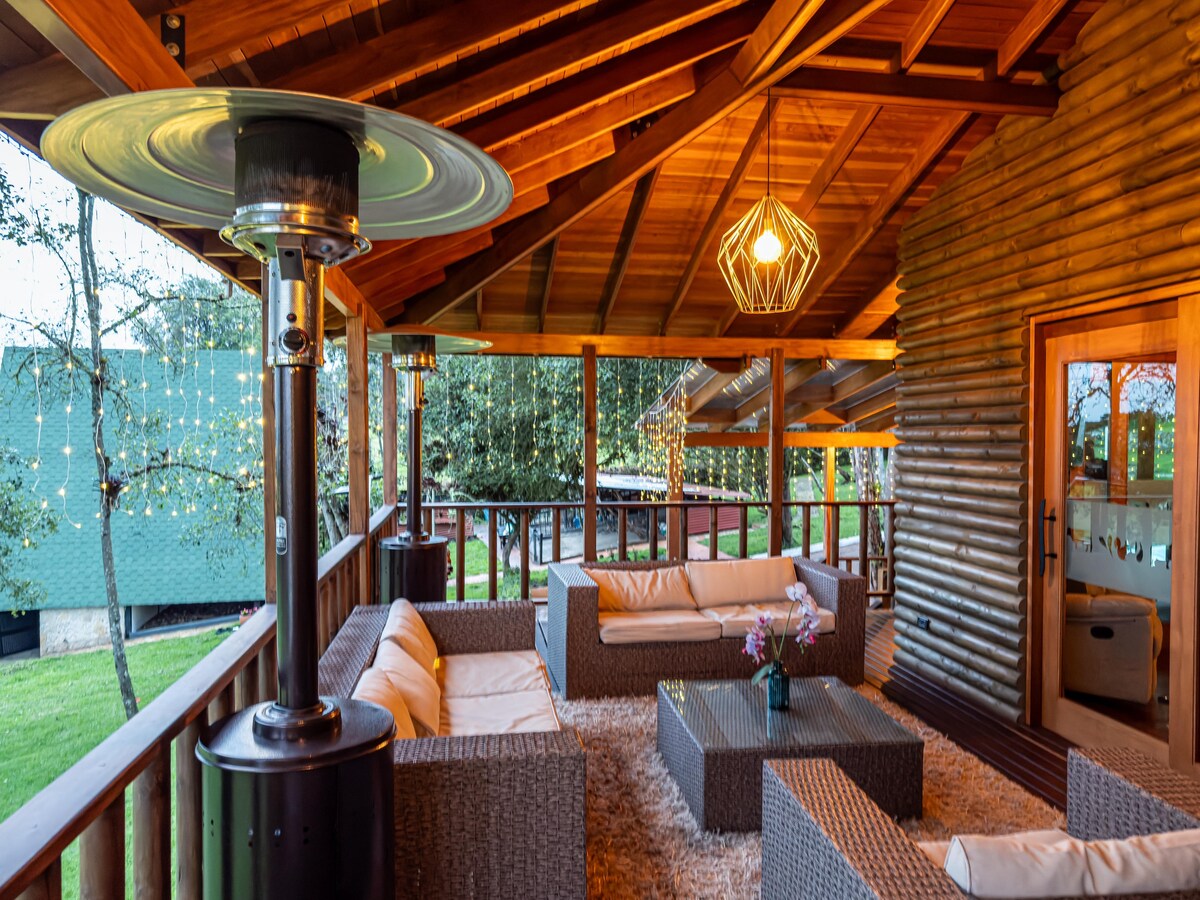 An outdoor seating area is featured under a wooden roof, with a stylish gas heater positioned at one side. Comfortable furnishings are arranged around a central table, while warm lighting creates a cozy ambiance. Green grass is visible outside the railing.