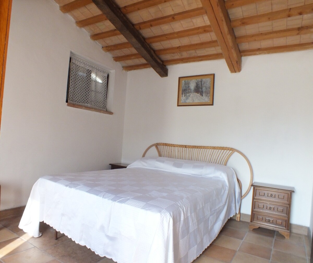A spacious sleeping area features a large bed with a white coverlet. Two wooden nightstands are positioned beside the bed, and a framed artwork hangs on the wall above. Exposed wooden beams stretch across the ceiling, while a window allows natural light to enter the room.