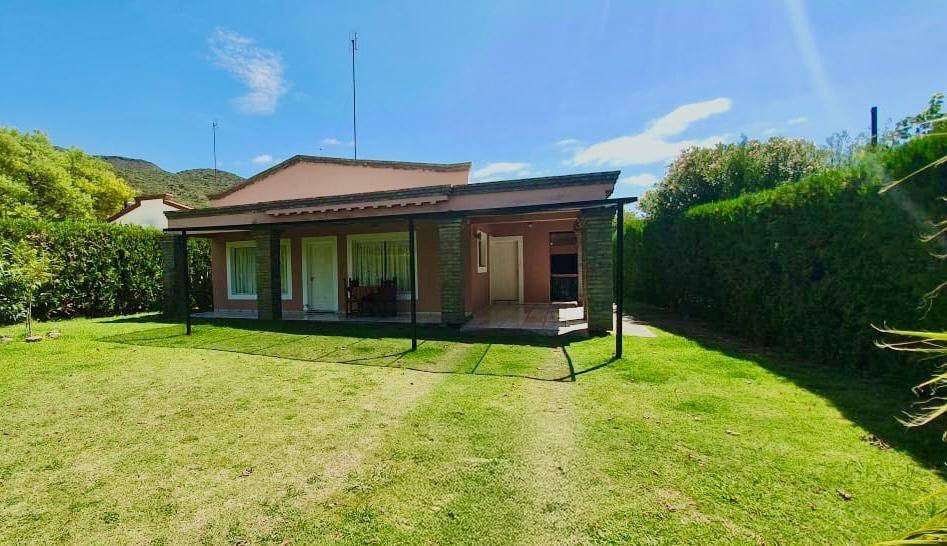 A single-story house is presented, featuring wide glass doors that open to a lush backyard. The exterior is framed by manicured hedges, providing privacy. A manicured lawn invites relaxation under clear blue skies, creating a serene outdoor space for enjoyment.