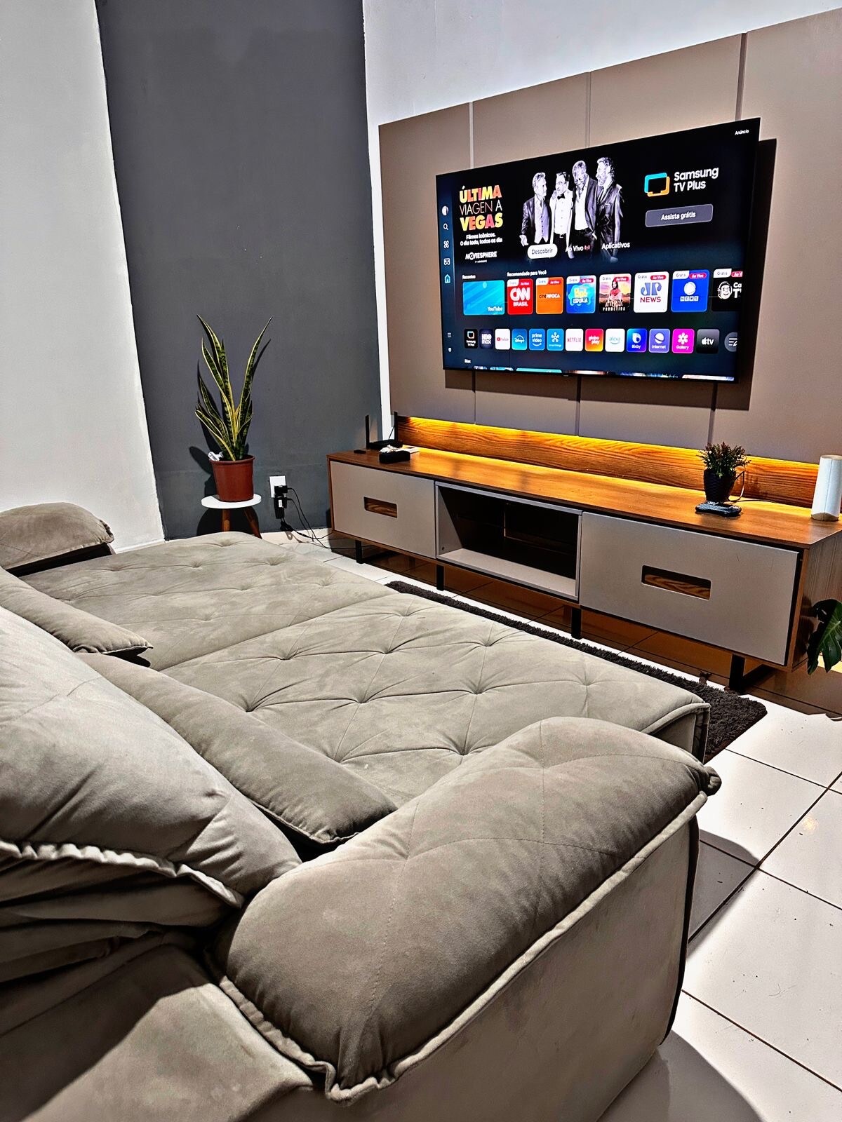 A comfortable seating area is shown, featuring a spacious, plush sectional sofa arranged opposite a mounted television. A modern sideboard beneath the TV displays decorative plants and offers storage. The wall is painted a muted tone, enhancing the area’s clean and contemporary aesthetic.