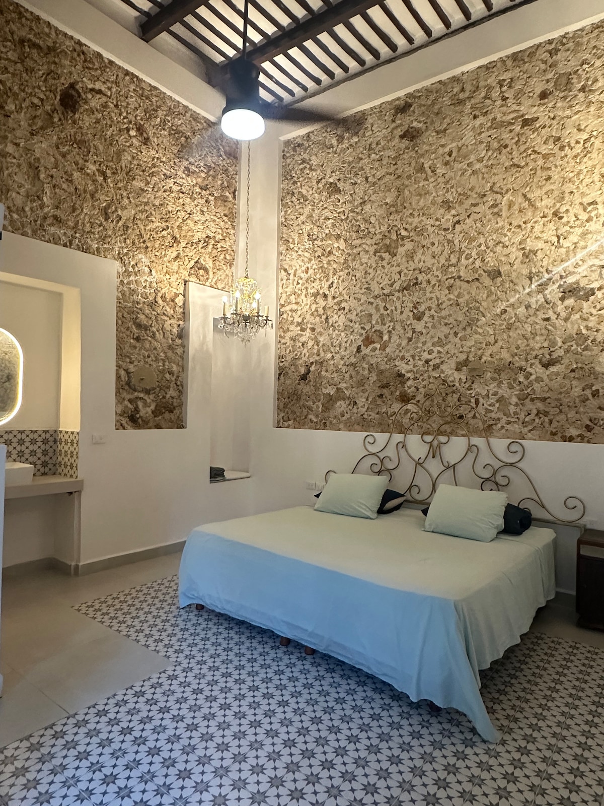 The sleeping area features a spacious bed with light bedding, complemented by two soft pillows. The textured, stone-like walls create an interesting backdrop, while patterned tiles adorn the floor. A chandelier offers ambient lighting, and a small window allows natural light to enter the room.