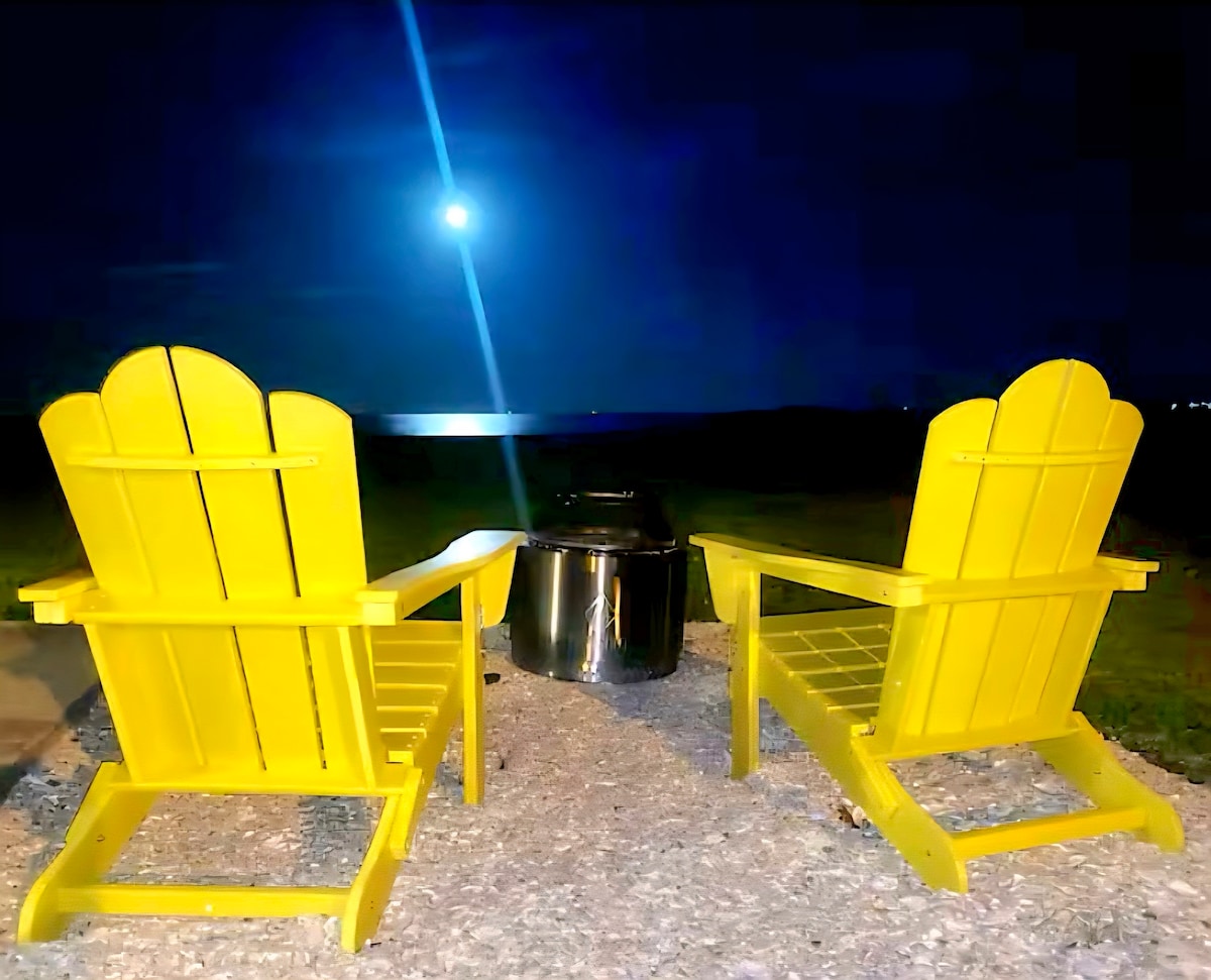 Two yellow Adirondack chairs are positioned facing a body of water under a night sky illuminated by the moon. A fire pit sits between the chairs, surrounded by gravel, providing a cozy setting for outdoor relaxation.