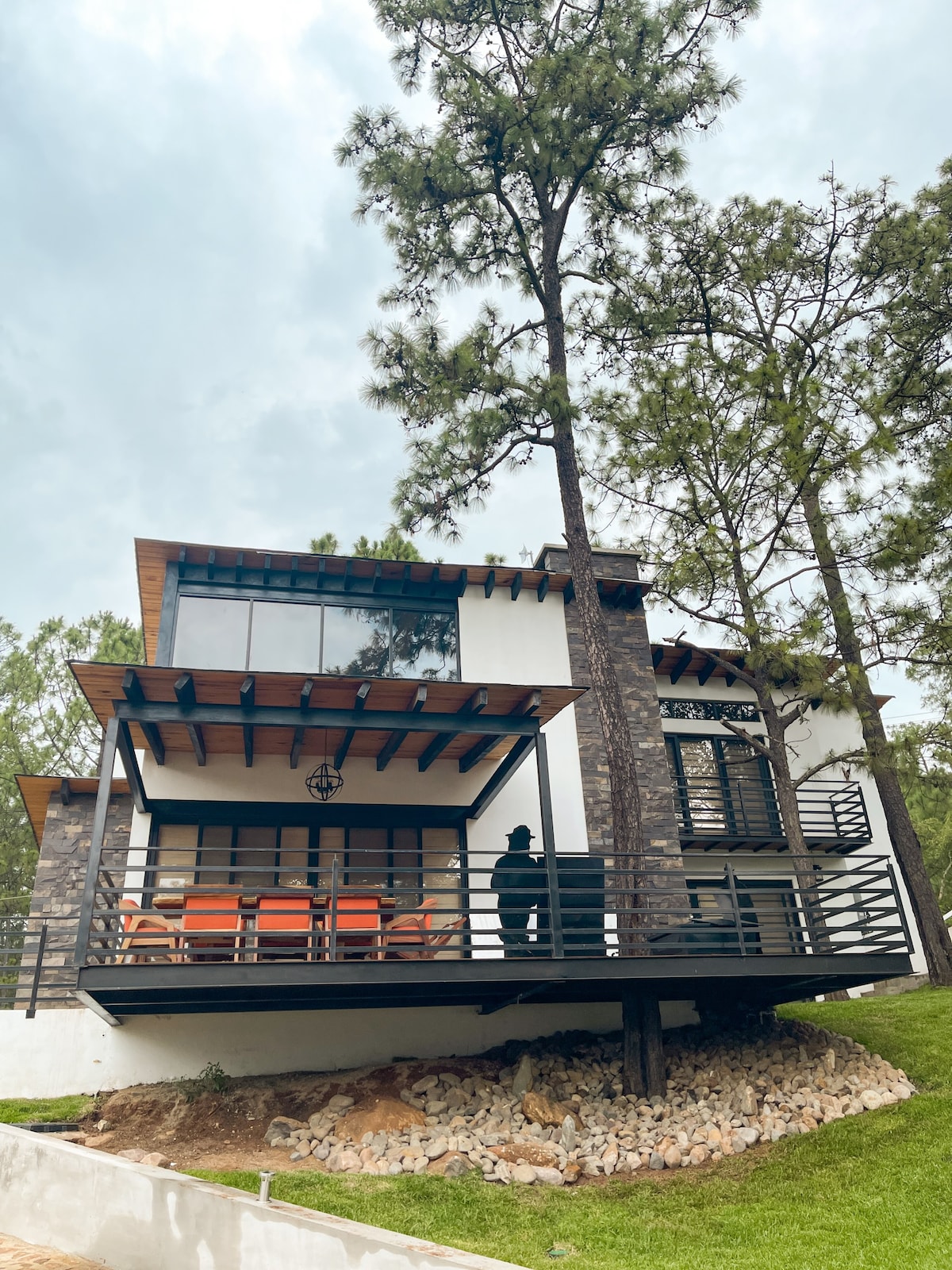 A modern house is set amid tall trees, featuring a balcony with orange chairs and a wooden ceiling. Large glass windows provide natural light, while stone accents add texture to the exterior. The landscaped surroundings include a grassy area and rocky elements beneath the structure.
