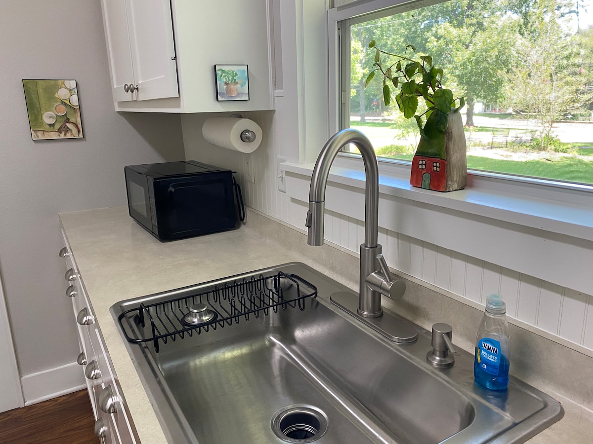 A clean kitchen area features a stainless steel sink beside a modern faucet. A compact microwave sits on the countertop, while a dish drying rack is positioned near the sink. Natural light enters through a window, showcasing a small plant and decorative item on the sill.