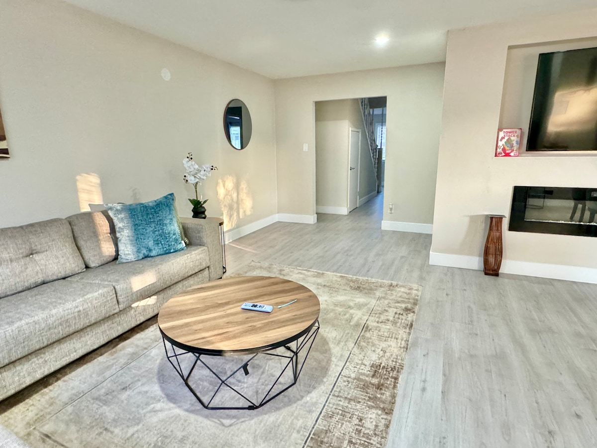 A spacious living area is highlighted by a comfortable gray sofa and a round wooden coffee table. Natural light filters through a large window, illuminating the light-colored flooring. A small contemporary fireplace is positioned against the wall, adding a subtle focal point to the room.