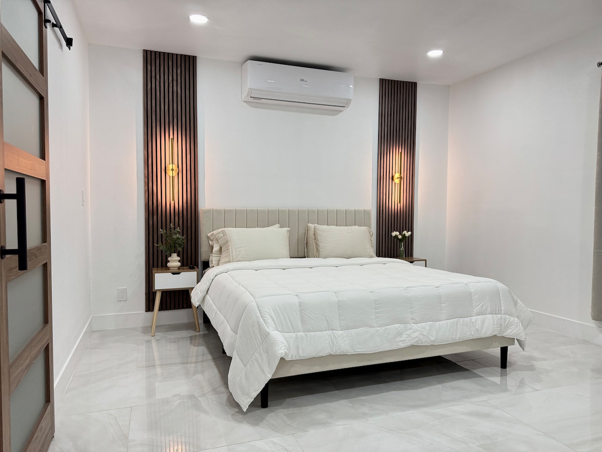 A spacious bedroom features a king-size bed with a soft, white comforter and two matching pillows. Accent lighting from the wall sconces adds a soft glow. Modern furnishings include bedside tables with small plants. A sleek air conditioning unit is mounted above, maintaining comfort.
