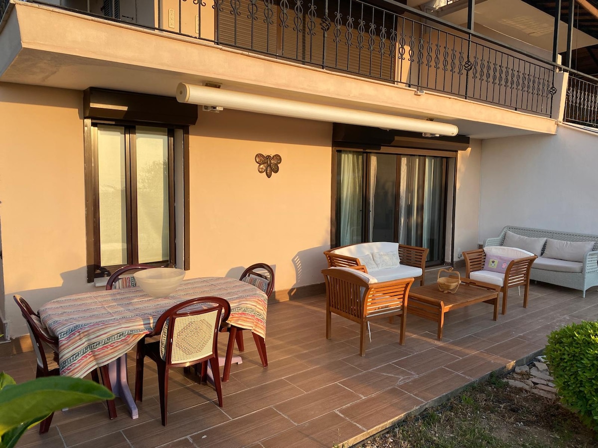 A covered outdoor terrace features a large table surrounded by four chairs, alongside a cozy seating area with a sofa and additional chairs. Large sliding doors allow for easy access, with natural light contributing to the inviting ambiance.