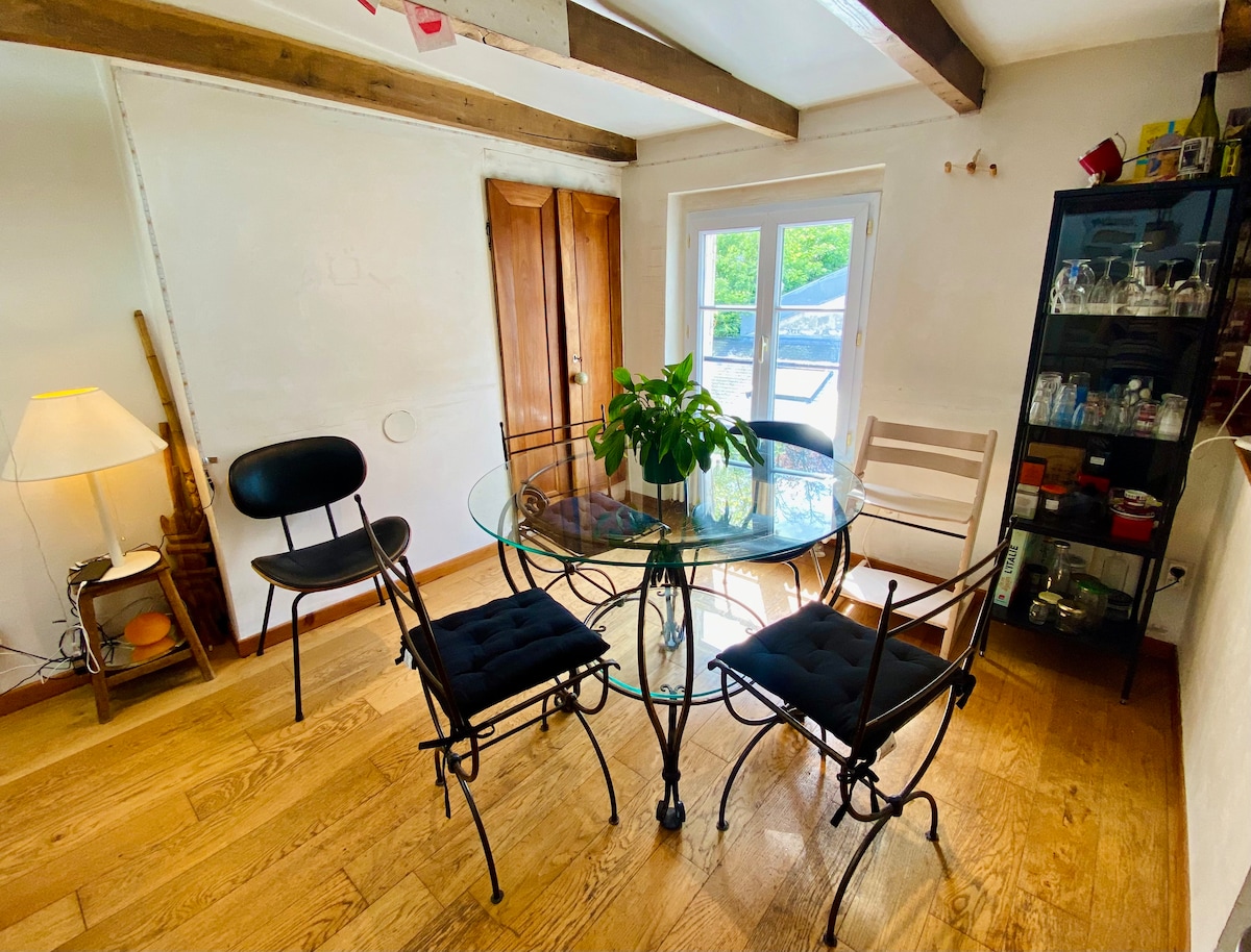 A dining area features a round glass table surrounded by four chairs, two with cushioned seats. Natural light enters through a set of double doors, leading to a view of the outdoor space. A small lamp and a glass cabinet display various items.