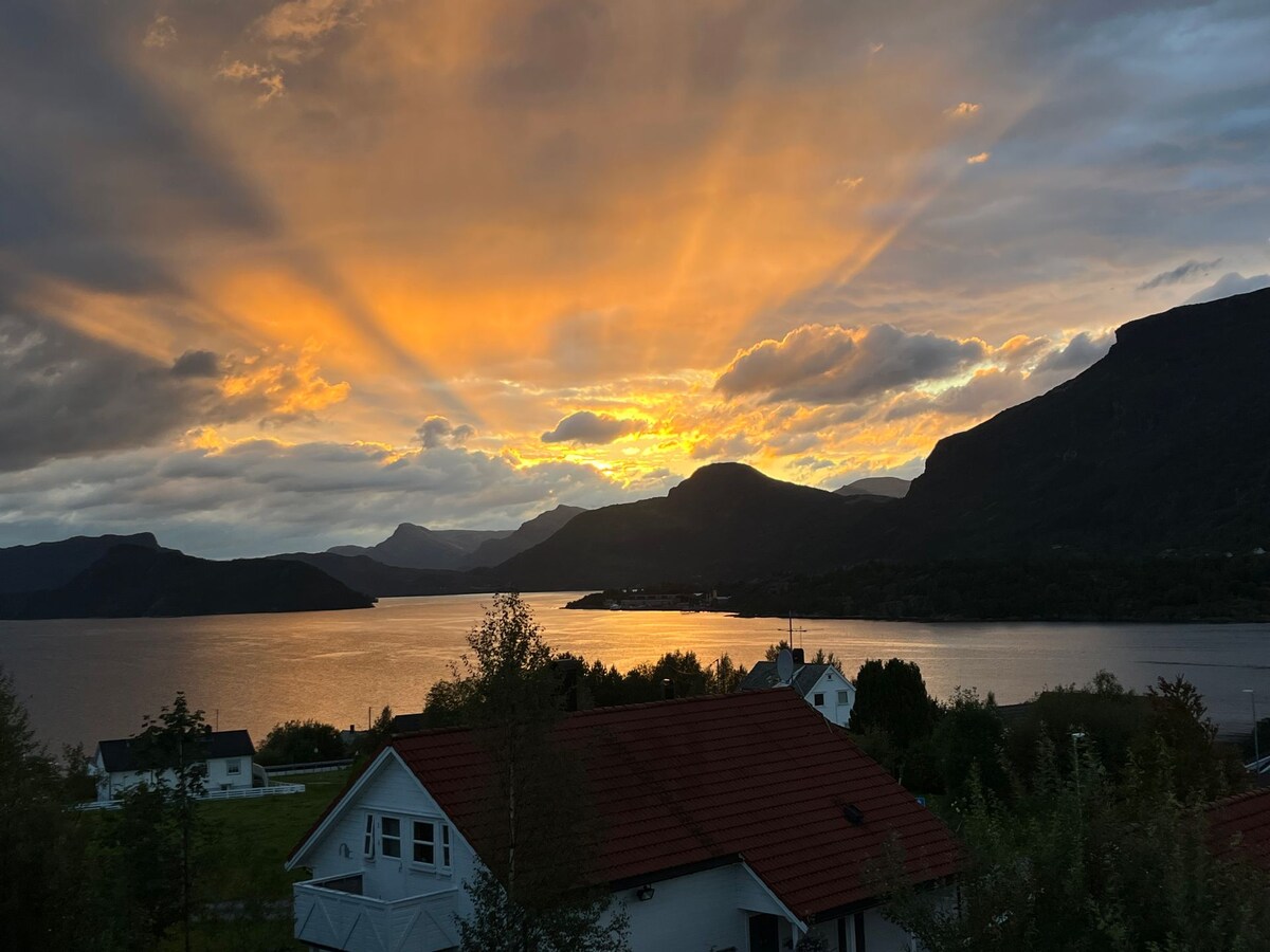 A stunning sunset displays vibrant rays of light across the sky, reflecting on the calm waters of the fjord. The surrounding mountains create an impressive backdrop, while the landscape features a mix of trees and houses, emphasizing the connection to nature.