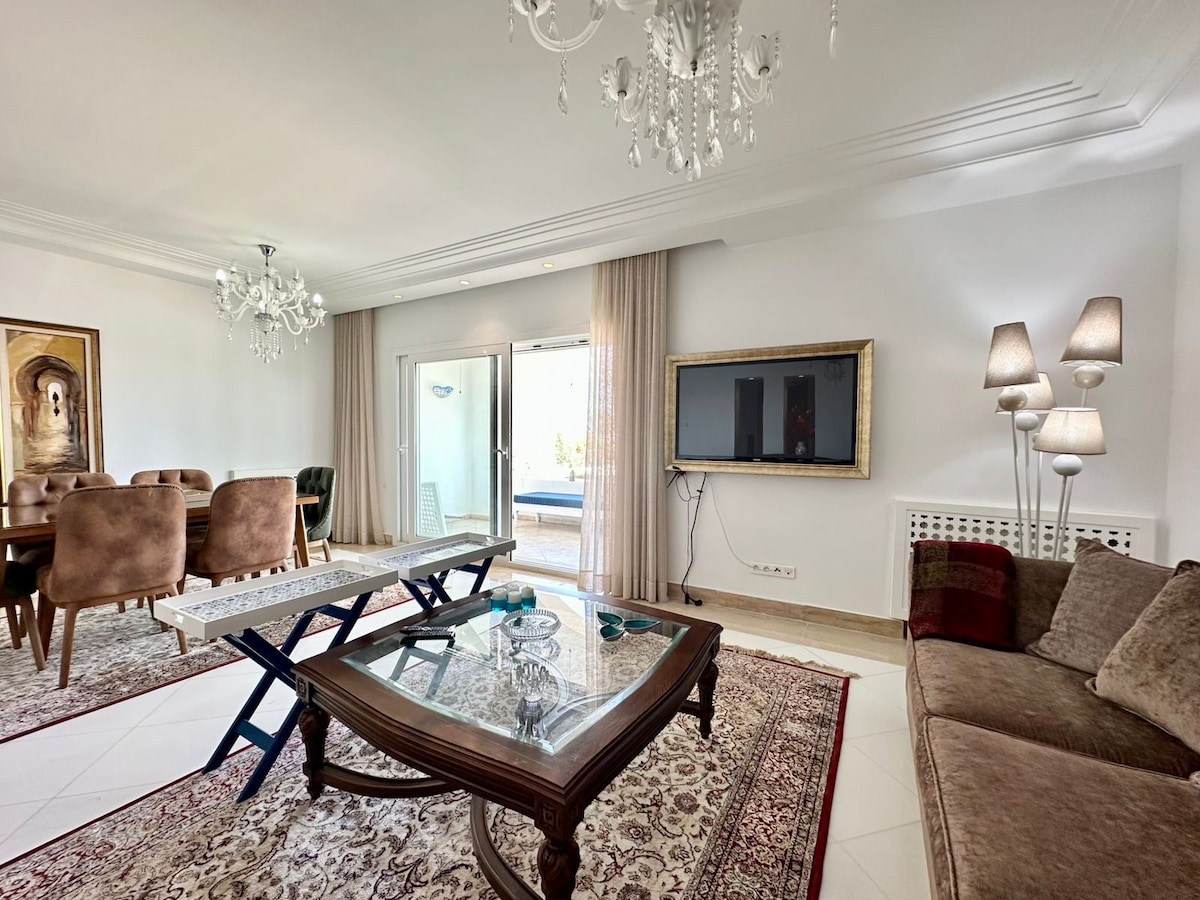 The living area is furnished with a combination of seating options including a plush sofa and stylish dining chairs. A glass table is positioned in the center, with decorative items displayed. Natural light enters through large sliding doors, which lead to an outdoor space.